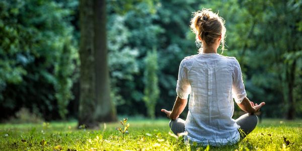 Young woman practicing mediation outdoors in a grassy field with trees surrounding the field.