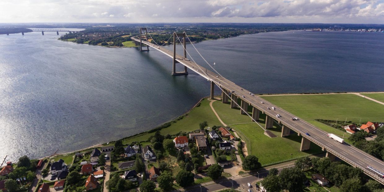 A large suspension bridge stretches over a wide river with houses nearby.
