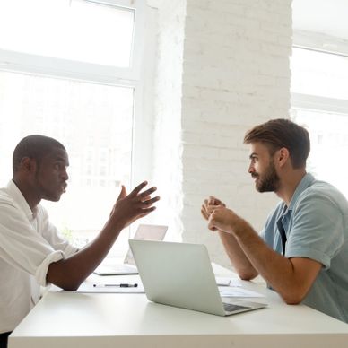 Two men engaged in a focused discussion over laptops in a bright office.