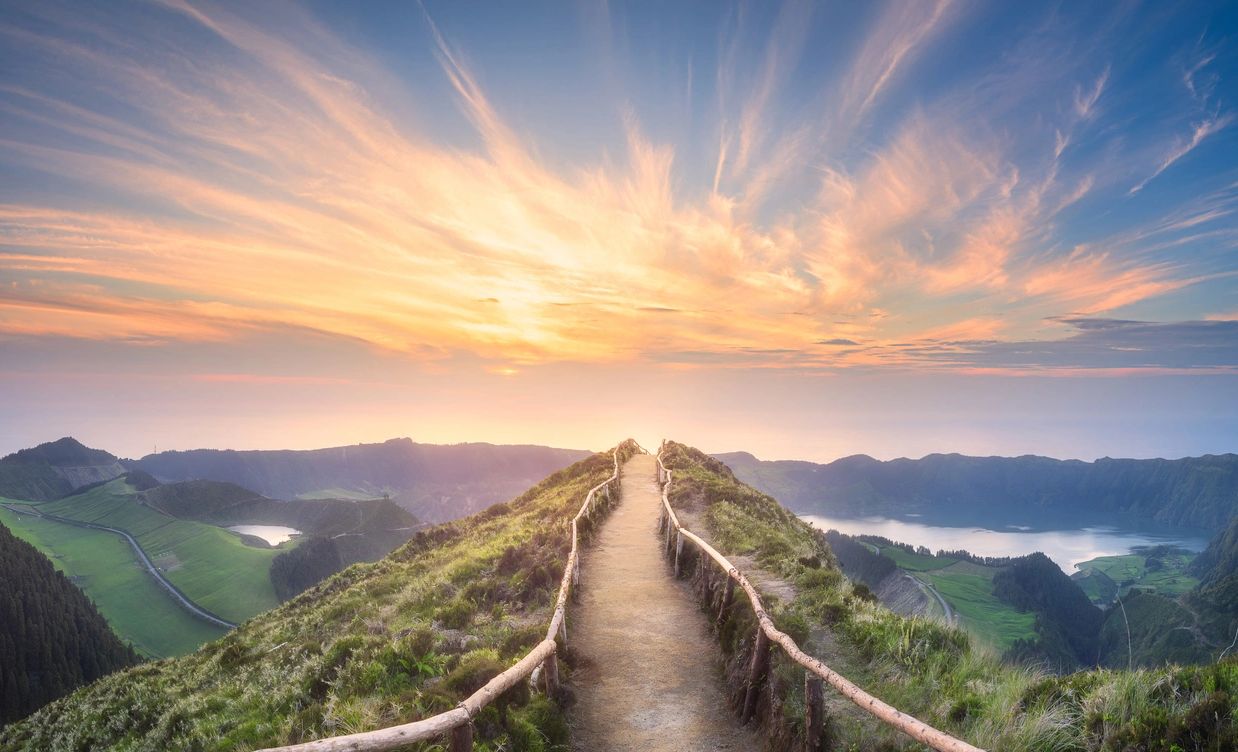 A sunset in wild looking clouds with a bridge across a mountain top leading to the sun