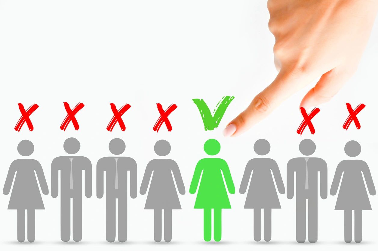 Male and female figurines in a single file line with red 'x's' above them and only one with a green checkmark above her.