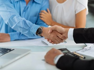 Two people shaking hands over a business meeting table with a couple in the background.