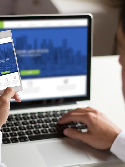 Person using a smartphone and laptop displaying a blue-themed website.