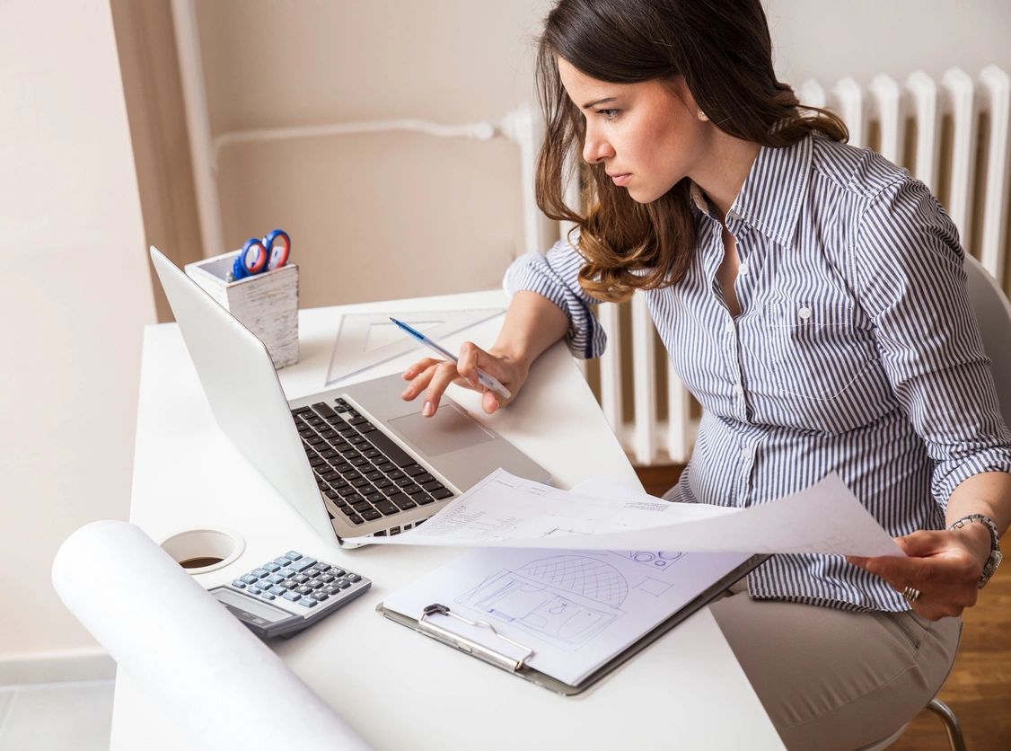A woman working on architectural plans with a laptop and documents.
