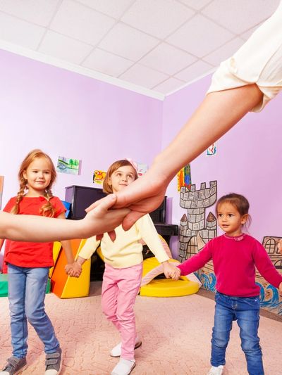 Children holding hands in a circle with an adult in a colorful playroom.