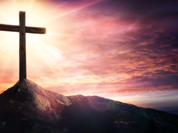 A wooden cross on a hill with radiant sunlight and a colorful sky.