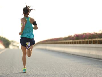 A woman in athletic wear and running shoes runs away from the camera on a paved road.