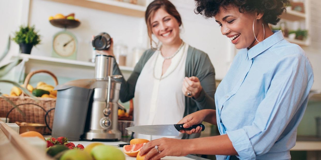 Two women happily prepare fresh ingredients in a kitchen, using a juicer & cutting fruits