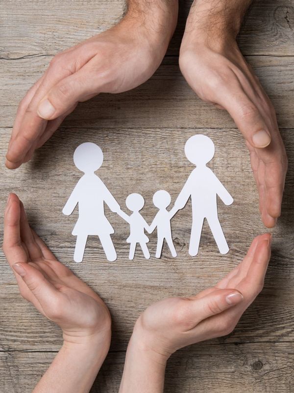 Hands protect paper cutout family on wooden surface.