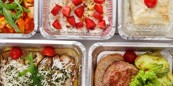 Assorted ready-to-eat meals in aluminum containers with fresh vegetables and toppings.