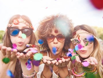 Three women blow colorful confetti towards the camera outdoors.