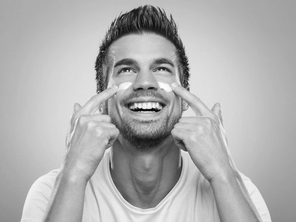 A man is smiling joyfully while applying a white face cream to his cheeks with his pointer fingers