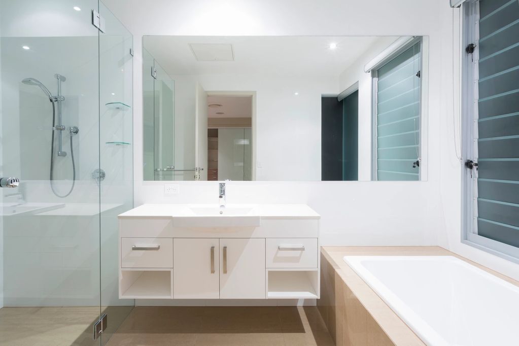 Modern bathroom with white vanity, large mirror, glass shower, and bathtub by window.