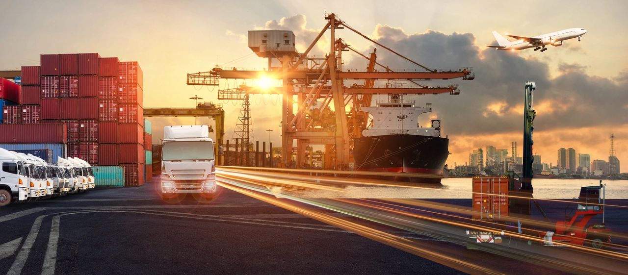A logistical hub shows parked tractors, stacked tractor trailers, one tractor driving forward, a ship vessel, an airplane and a forklift right off the water.