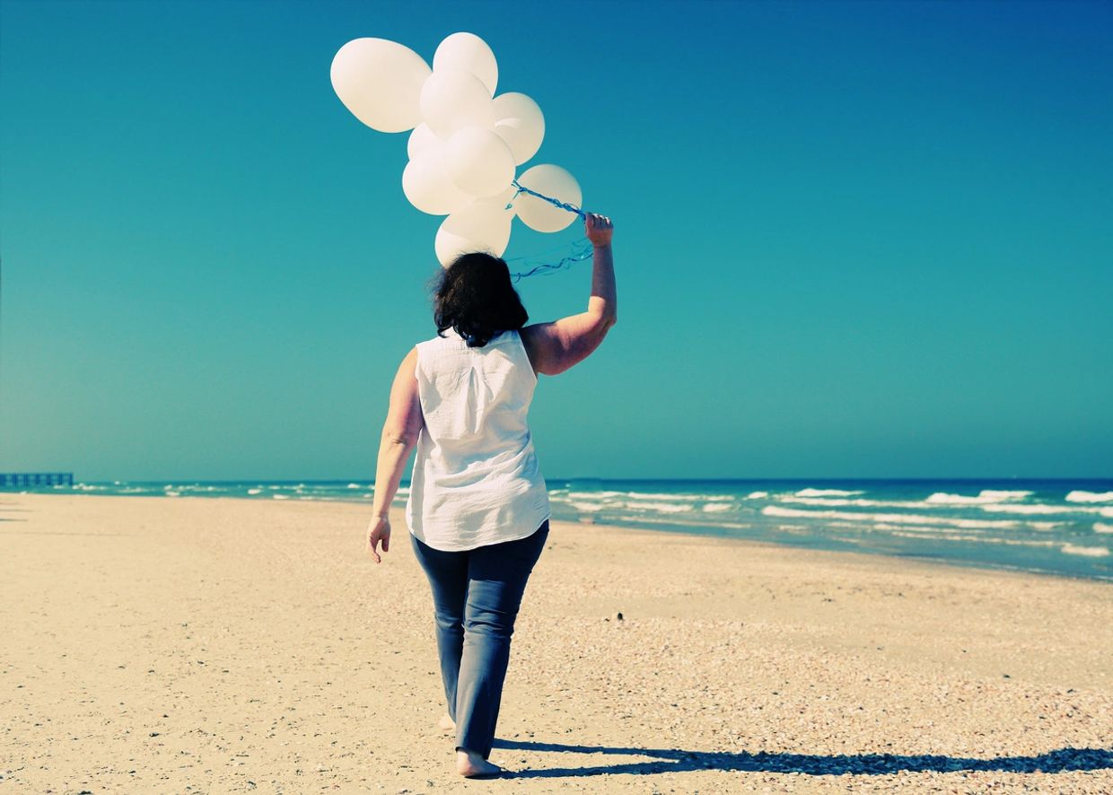 Woman holding white balloons walks on a secluded beach.