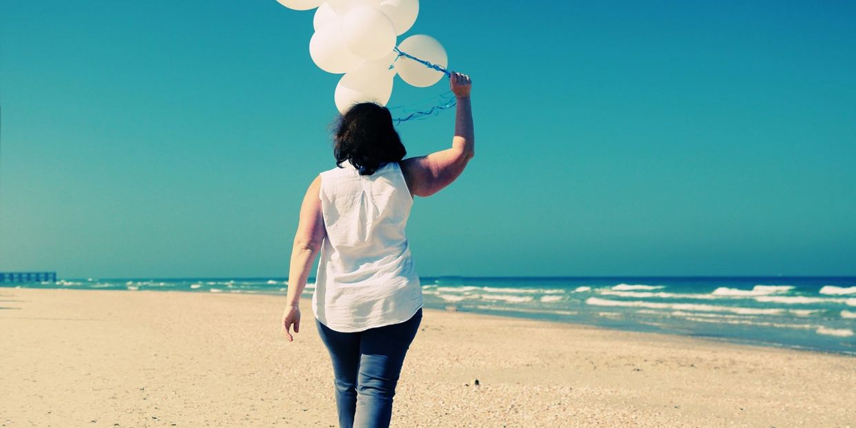 Woman holding white balloons walking on a sunny beach.