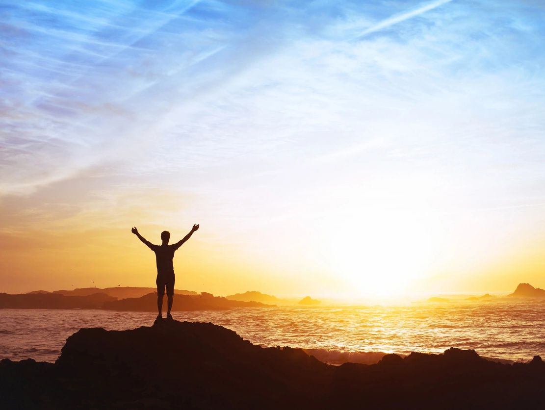 A person stands with arms raised on a rock at sunset by the ocean.