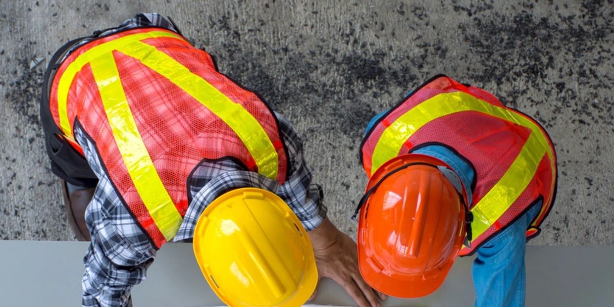 Two construction workers in safety vests and helmets examining plans.