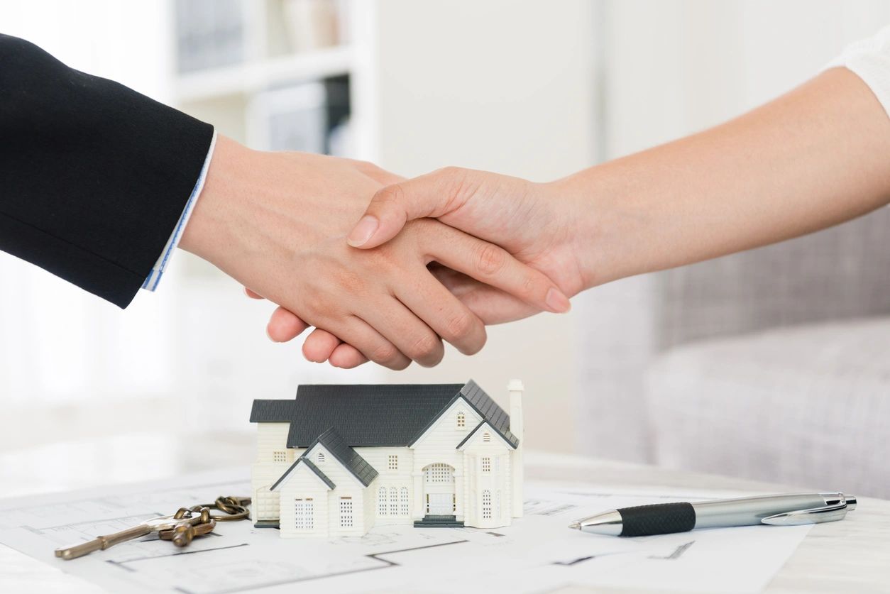 A buyer and seller shake hands above a house figurine and the home's blueprint.