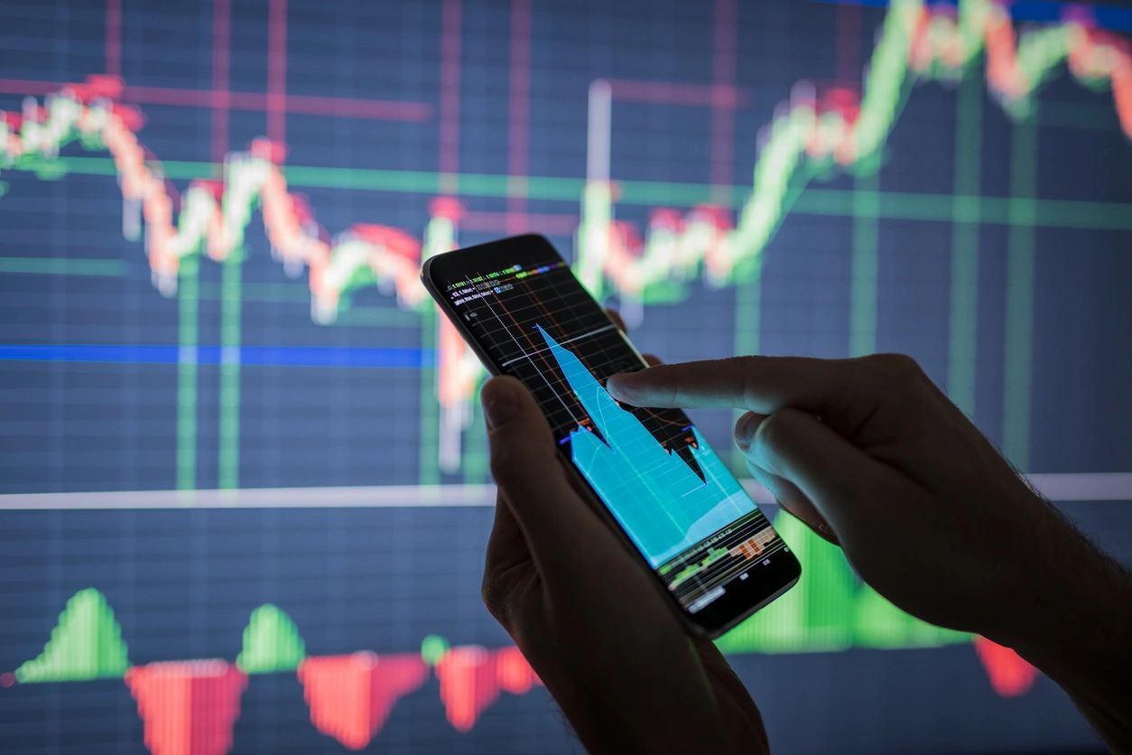 A person points out a point on the stock chart on their cell phone with another monitor in front of them with another stock chart.