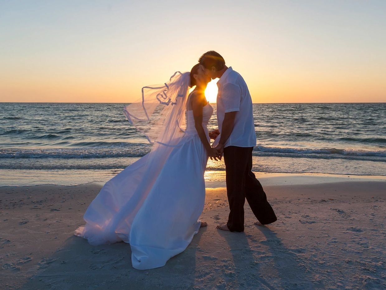 Newlyweds share a kiss on the beach at sunset.