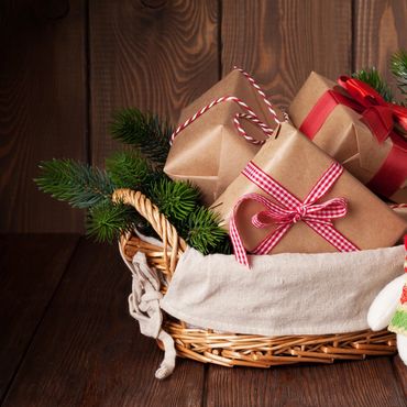 Wicker basket with wrapped Christmas gifts and pine branches on wooden floor.
