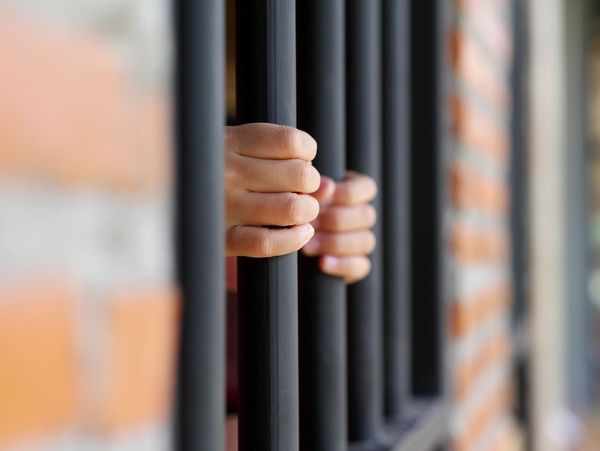 Hands gripping black prison bars behind a brick wall.