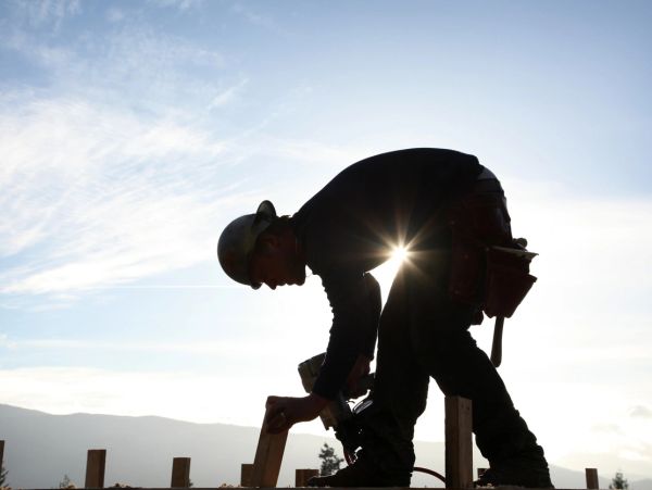 Silhouetted construction worker using a nail gun under a bright sky.