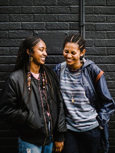 A picture of 2 black women laughing with each other, standing against a black brick wall
