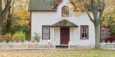 Charming white house with a red door surrounded by autumn foliage and a white picket fence.