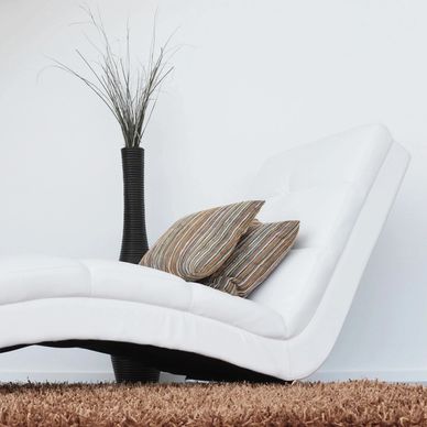 Modern white chaise lounge with striped pillows and a tall black vase on a brown rug.