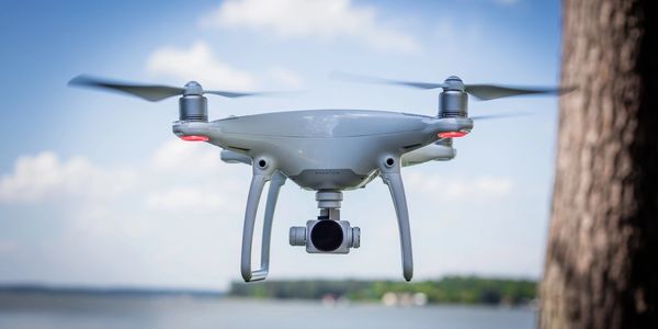 A white quadcopter drone flying outdoors near a tree and lake.