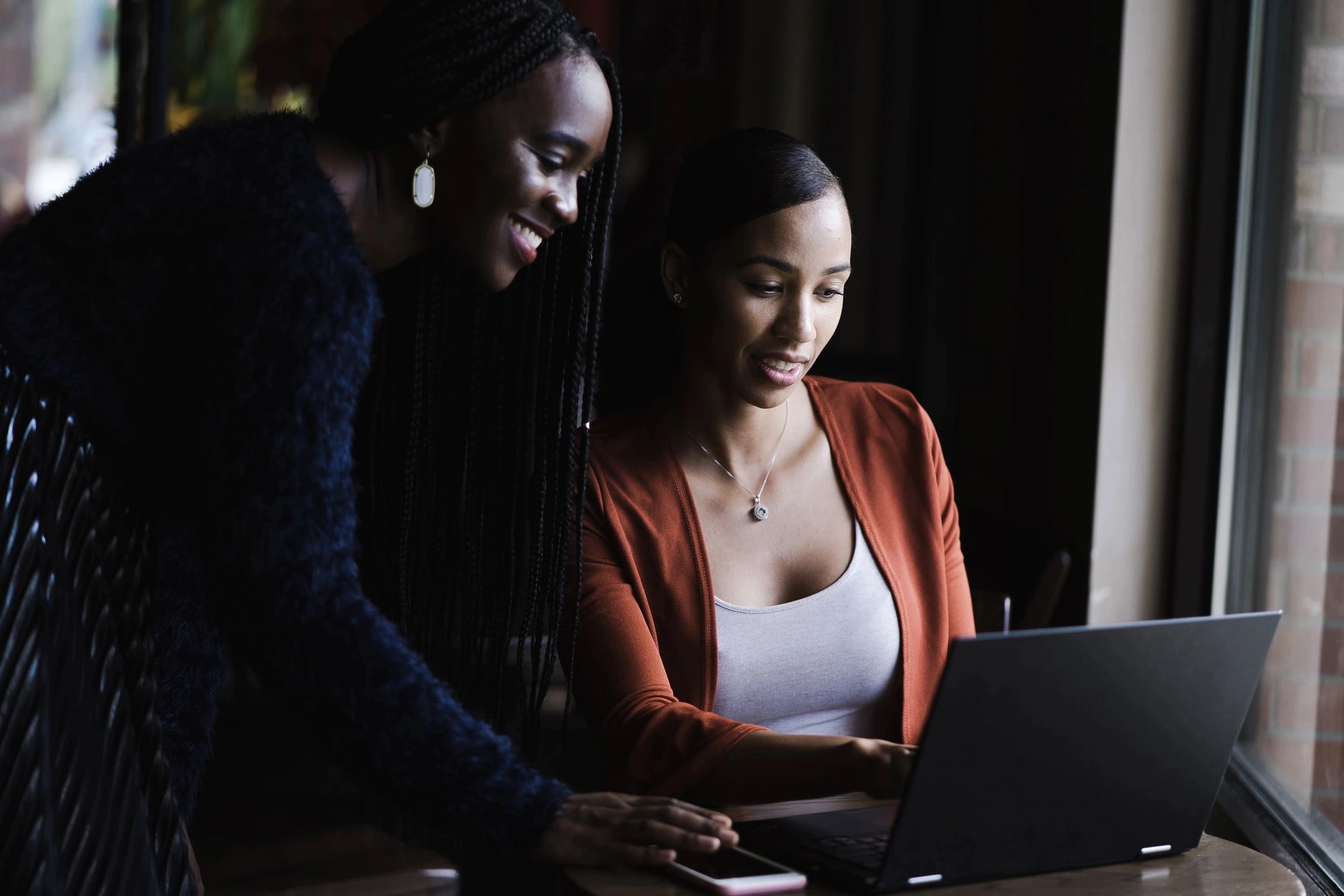 Two smiling women sitting at a table are sharing information on a single laptop computer screen.