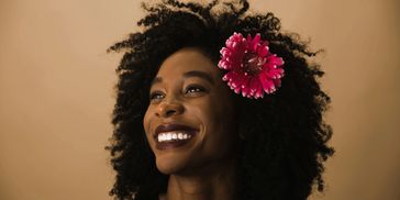 A woman smiles brightly with a pink flower in her curly hair against a beige background.