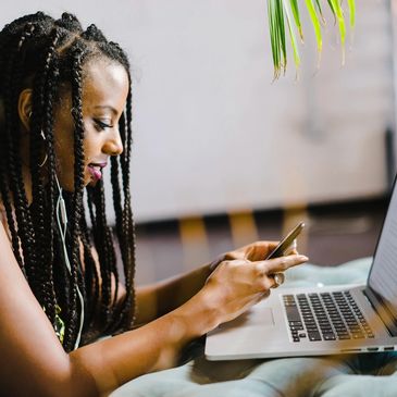 A woman with braids sitting and sending a text message with her laptop in front of her.