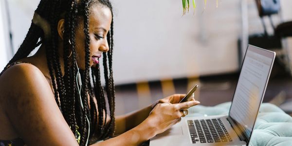 Black women sitting in her chair, while on her phone in front of a computer. 