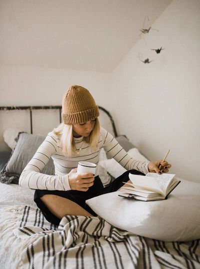 A woman sitting on a bed, coffee in hand, while writing in a journal.