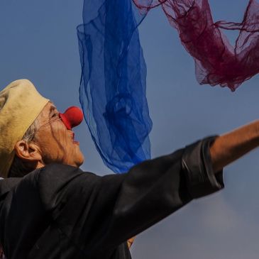 Elderly person with a red clown nose and beret tossing colorful scarves in the air.