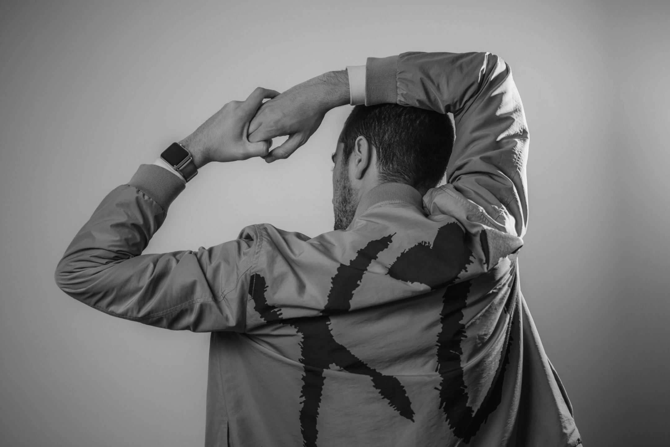 Man stretching with a brown jacket and smartwatch, back facing the camera.