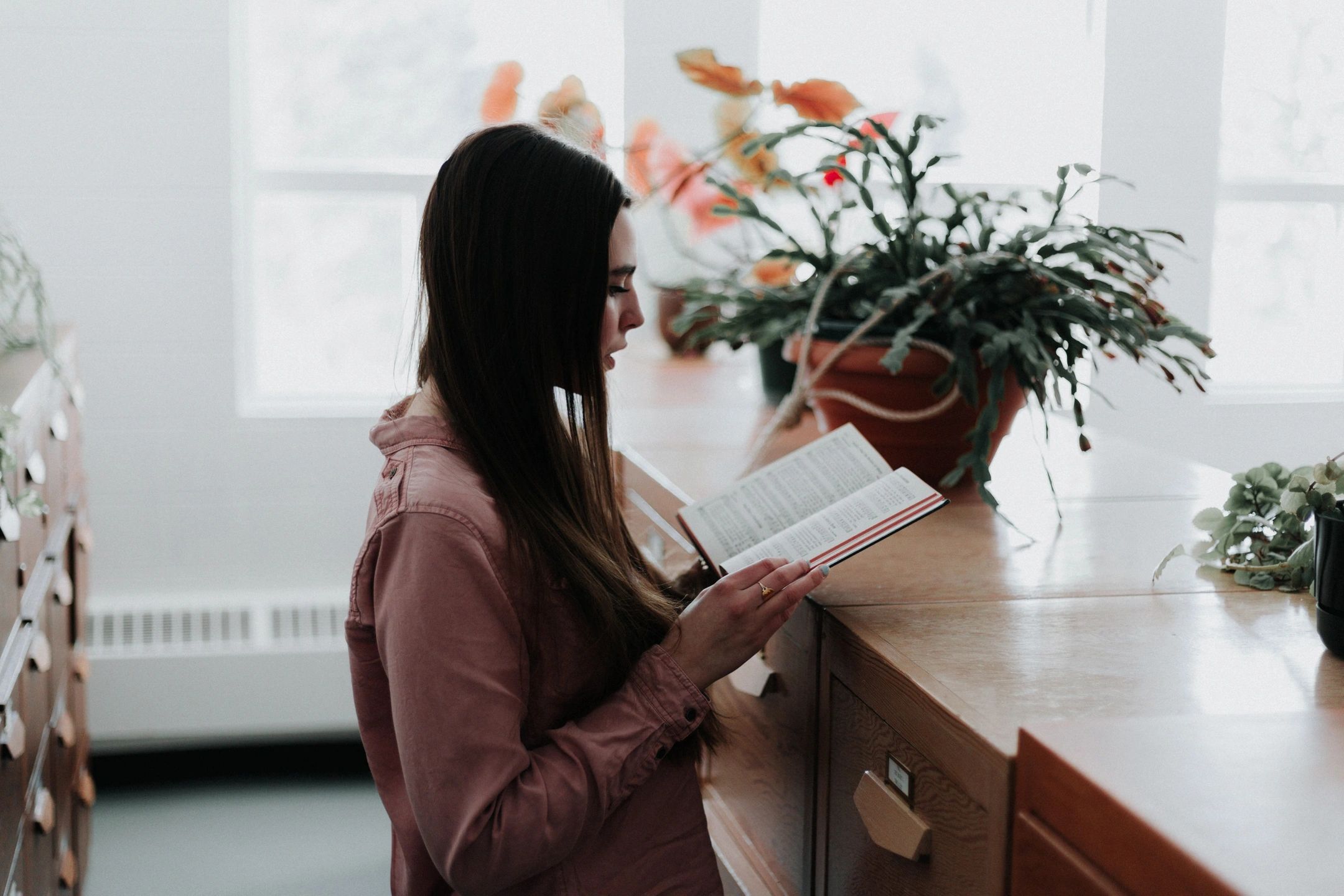 A woman with long hair reads a book beside a wooden table with potted plants