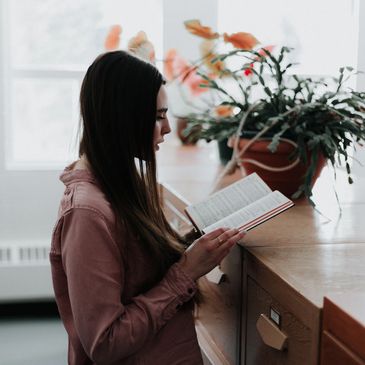 A woman reads a book near a window and potted plant indoors.
