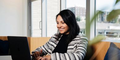 Seated woman with dark hair in striped sweater smiles down at laptop in front of her