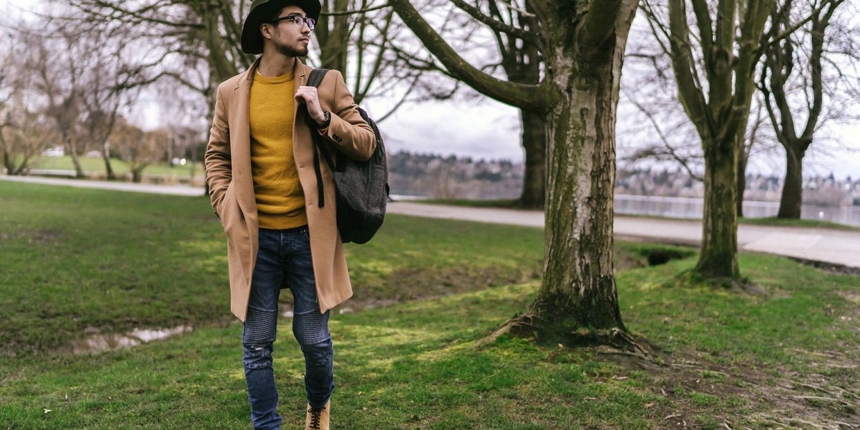 Man in a camel coat and hat walking in a park with a backpack.