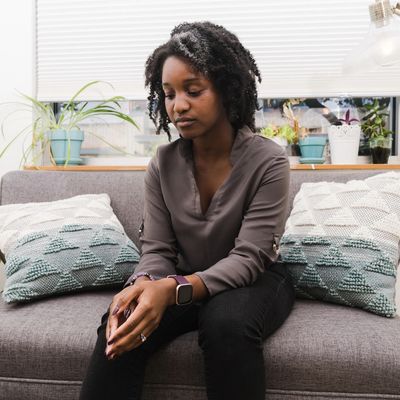 A black female sitting on a coach with her hands clasped looking down. 