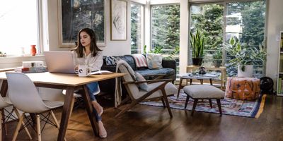 A woman working on a laptop in a cozy, bright living room with large windows.