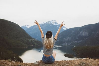 girl sitting on a rock facing a mountain and lake landscape, making "rock and roll" hand gesture