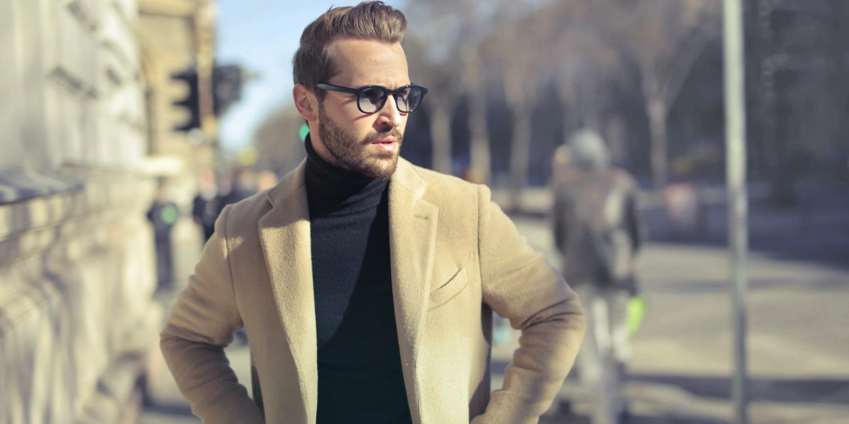 Man in beige coat and black turtleneck standing outdoors, looking to the side.