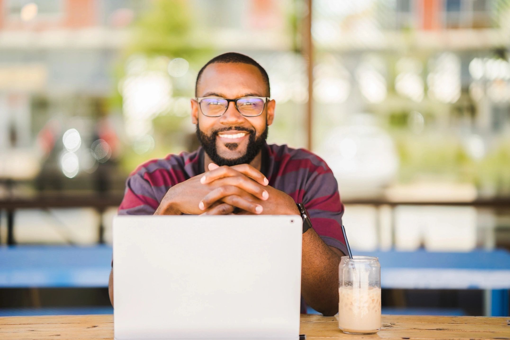 The man sat at a desk looking at the camera with a big smile and his computer is open on the desk.