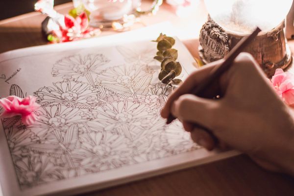 A person sketches detailed flowers in a coloring book with natural light.