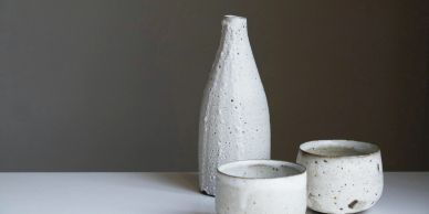 Minimalist ceramic bottle and cups with a rustic white glaze on a white surface.